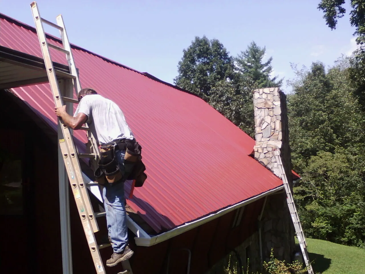 Roofer on a ladder beside a red metal roof and stone chimney — Soffit & Fascia Repair by Roofing Brothers in Gouldsboro